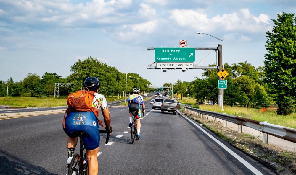 Two cyclists ride on a road with cars, approaching a sign indicating the Belt Parkway towards Kennedy Airport. The sky is partly cloudy and the surrounding area is green with trees.
