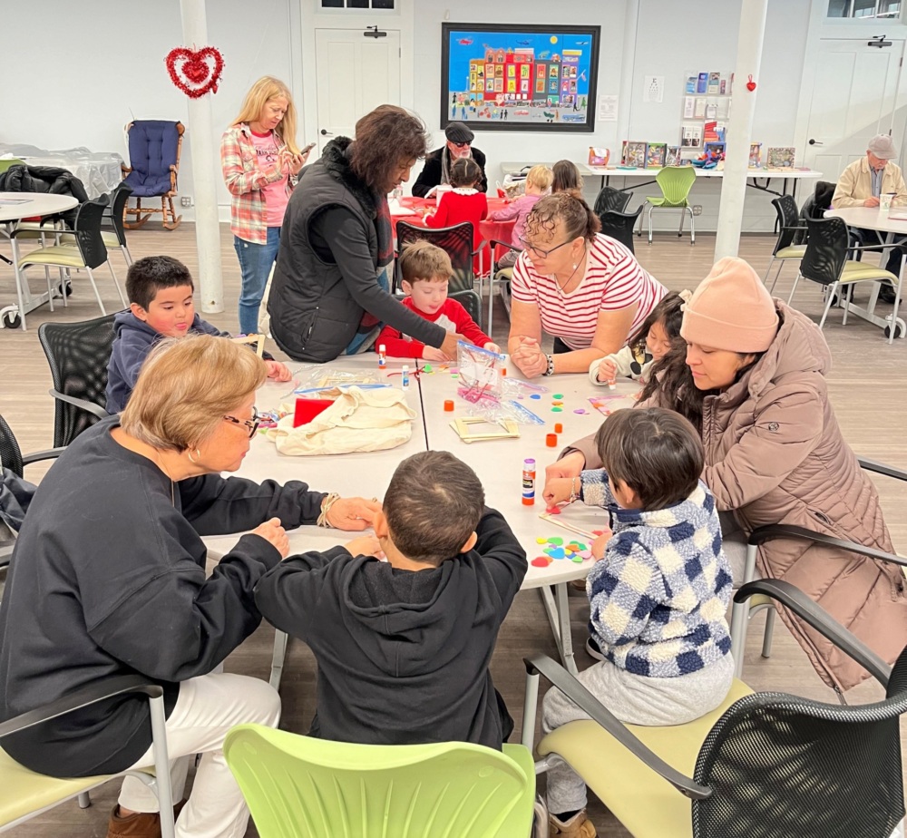 Several adults and children sit at tables in a classroom, engaging in various arts and crafts activities. A bulletin board with colorful papers is visible in the background.