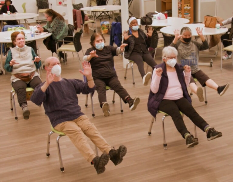 A group of older adults seated on chairs participate in an exercise class. All participants are wearing masks and are lifting their arms and legs. Other people are seen in the background.