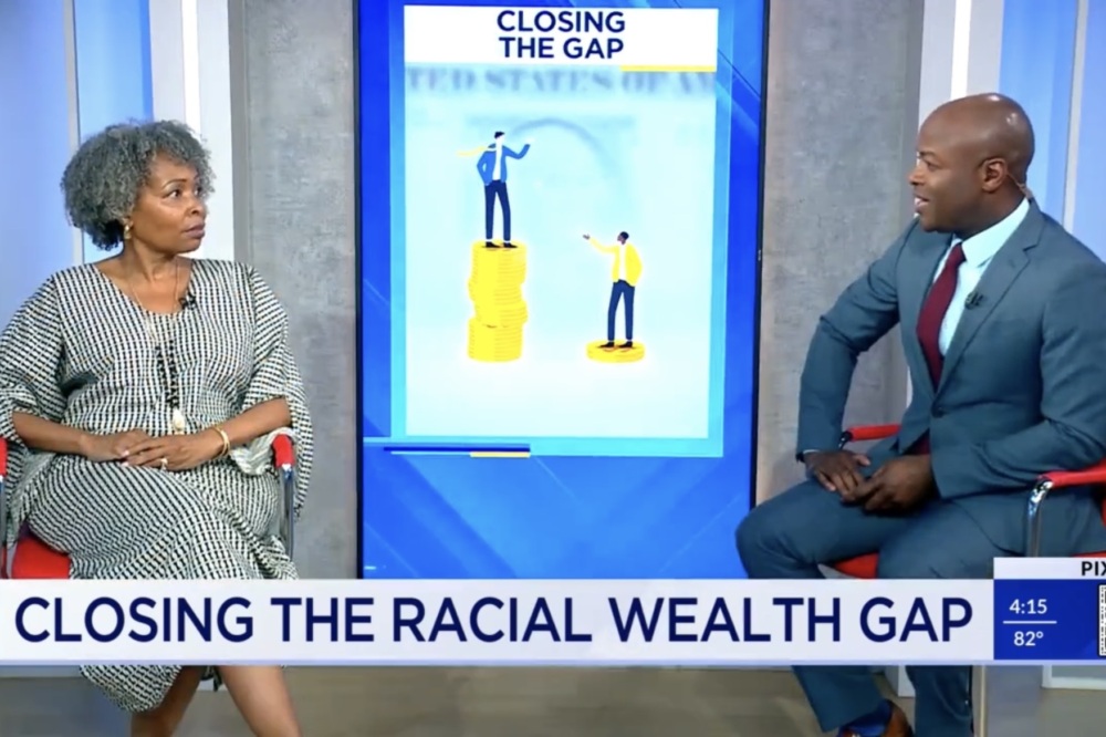 Two people seated in a studio discuss "Closing the Racial Wealth Gap," with a graphic of uneven coin stacks and titled "CLOSING THE GAP" displayed on the screen behind them.