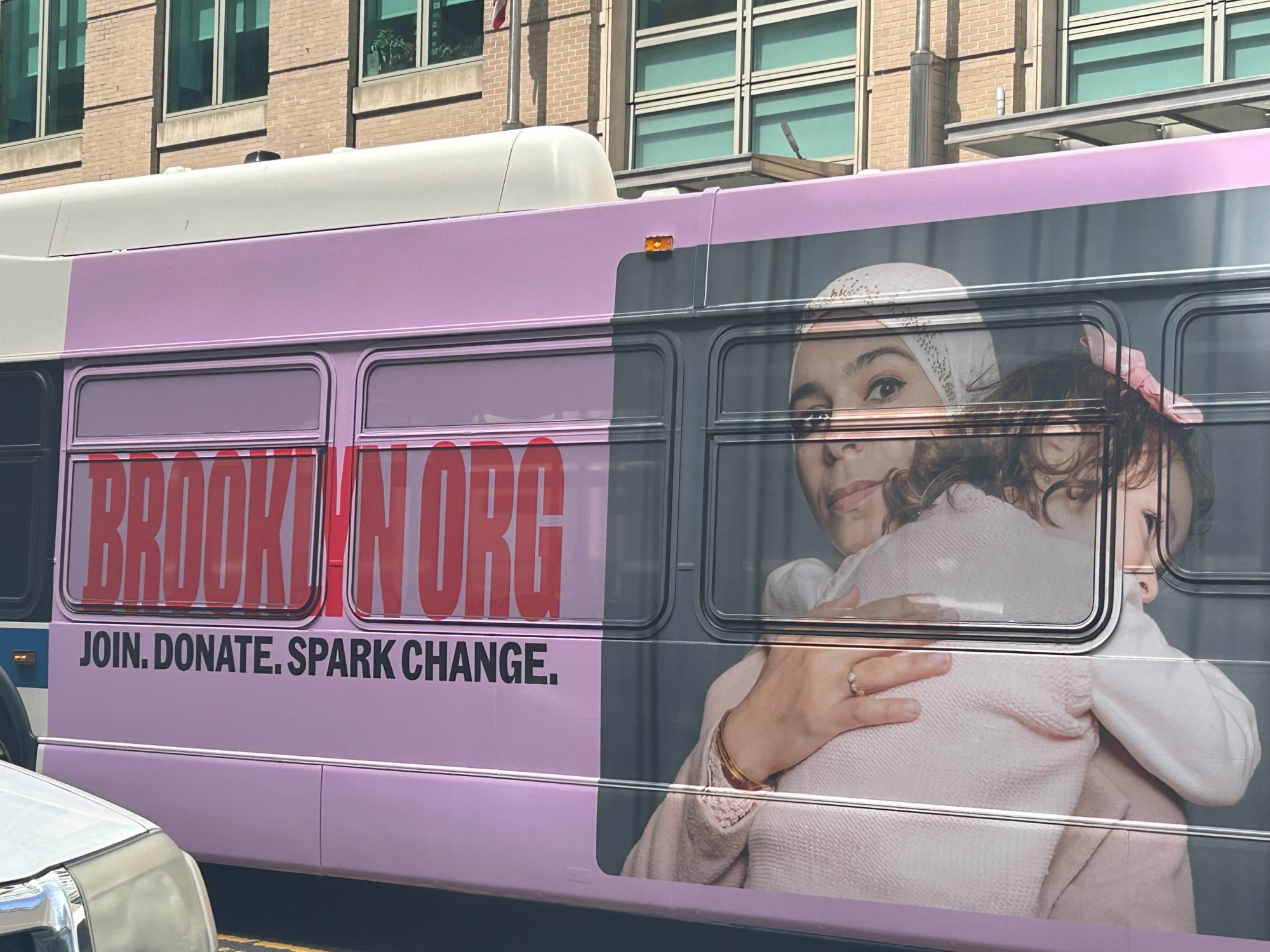 A bus with an advertisement side shows a woman holding a child. The text reads "BROOKLYN.ORG. Join. Donate. Spark Change." It's a heartfelt call to action, encouraging everyone to give to Brooklyn and make a difference in the community.