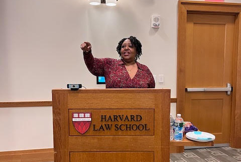 A person stands at a podium with the Harvard Law School logo, pointing while speaking. The setting appears to be a lecture or presentation room.