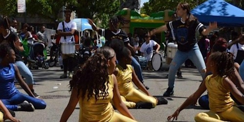 A group of dancers in gold and blue outfits performs in a street festival with onlookers and drummers in the background, under sunny weather.