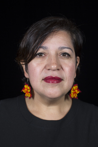 A woman with short dark hair wearing red and yellow flower earrings and a black top against a black background.