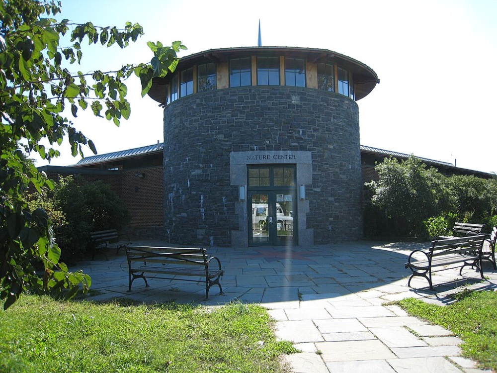 Circular stone building with a sign reading "Nature Center" above the entrance, surrounded by greenery and benches on a sunny day.