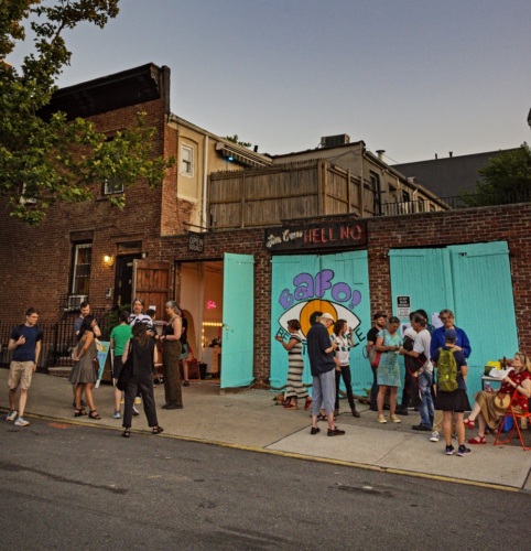 A group of people gather outside a brick building with mural art under a clear evening sky with a visible half-moon.