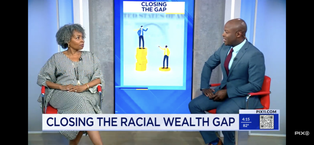 Two individuals, a woman in a checked dress and a man in a suit, are seated and discussing "Closing the Racial Wealth Gap" as displayed on a screen between them on a PIX11TV news segment.