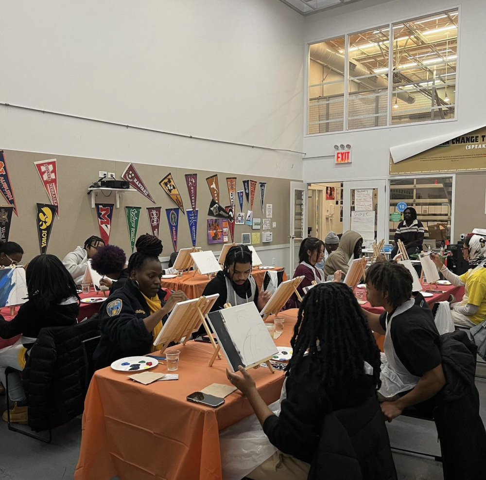 A group of people sit around tables in a brightly-lit room, each working on individual canvases with paint. Various college pennants decorate the walls, and supplies are scattered on the tables.