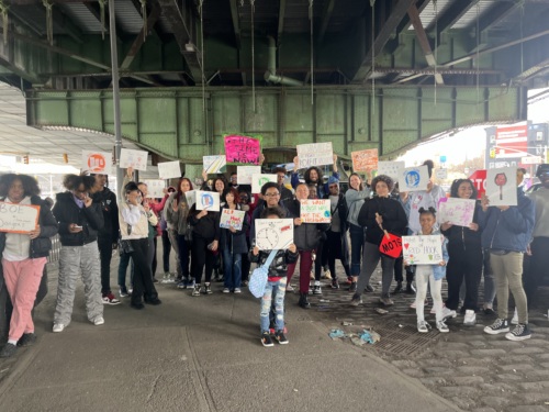 A group of people stand under a bridge holding various signs with messages. A child is in the foreground holding a sign with a clock drawn on it.