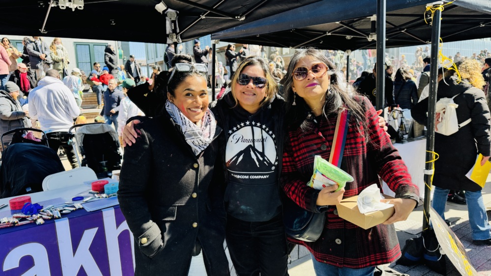 Three individuals pose together outdoors at a crowded event under a tent. They are dressed in casual, warm clothing; one of them holds food. A table with items and a sign is partially visible behind them.