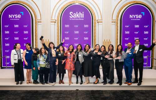 A group of people standing on a stage in front of NYSE and Sakhi for South Asian Women branded screens, posing for a photo, some with hands raised and smiling.