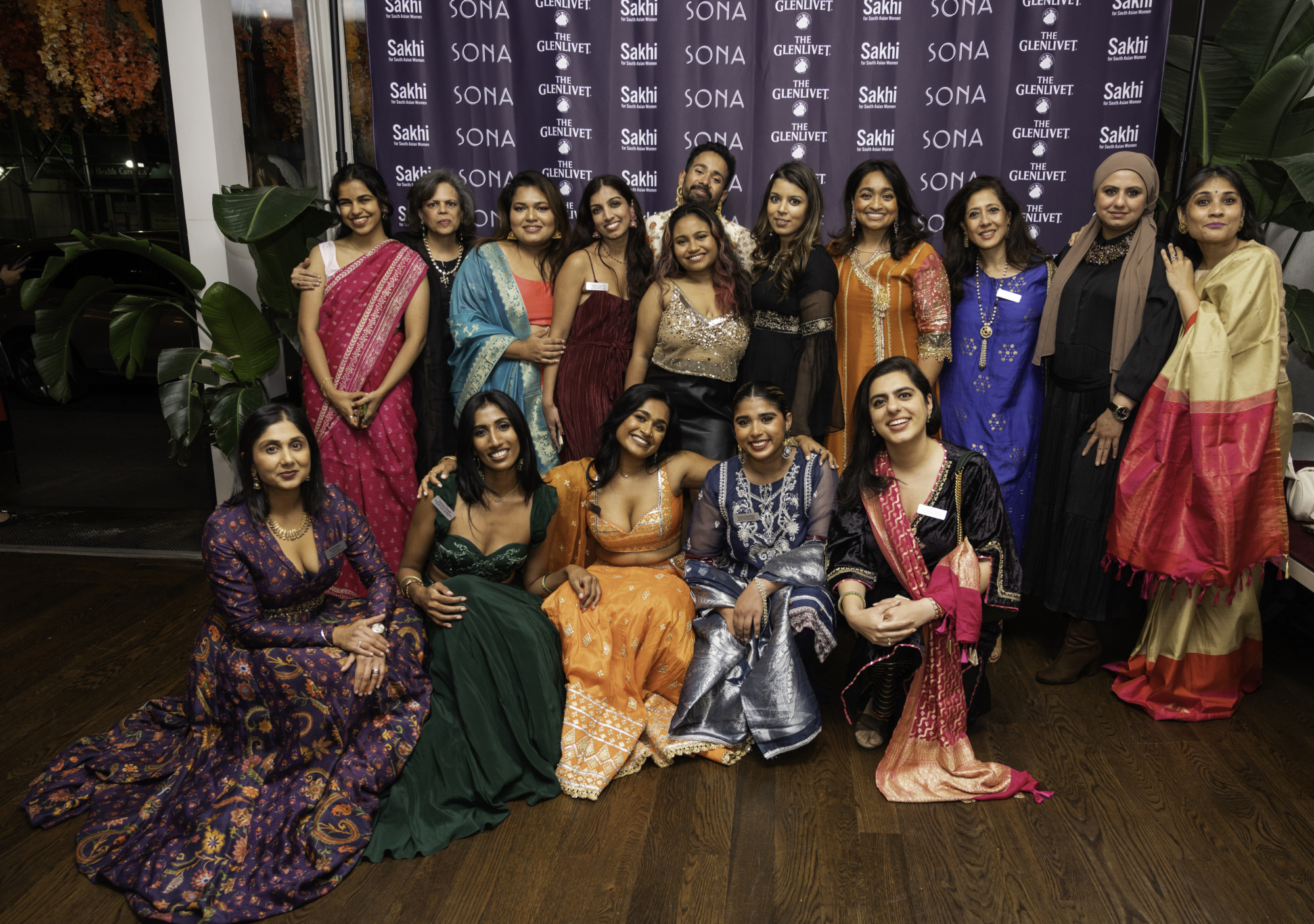 A group of women in colorful traditional attire pose together in front of a SONA branded backdrop. They are smiling and appear to be at an event.