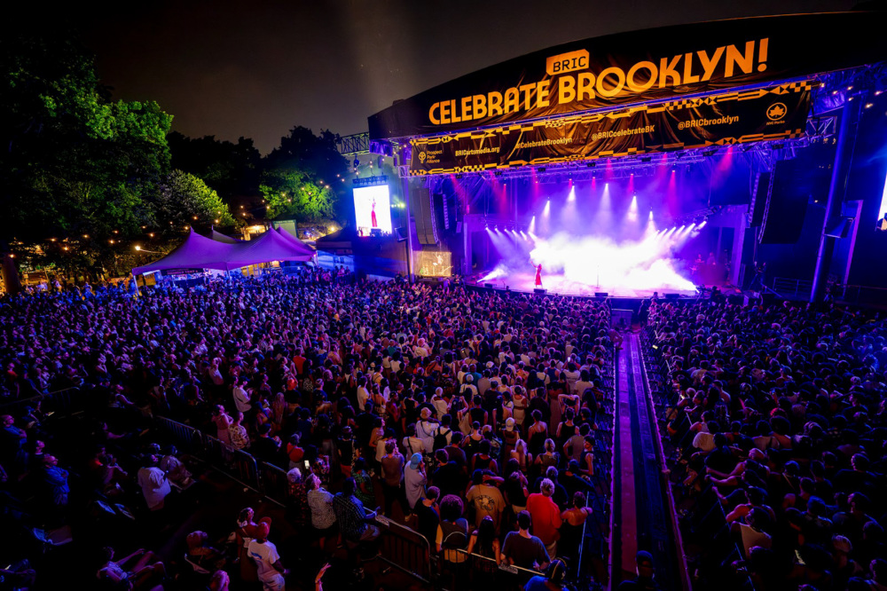 A large crowd gathers at an outdoor nighttime concert under a stage with a sign that reads "Celebrate Brooklyn!" The stage is illuminated with vibrant purple lights, and a performer is visible.