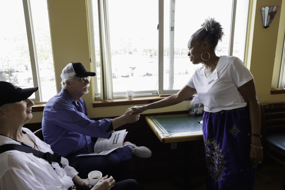 A woman in a white shirt shakes hands with a seated man in a blue shirt and cap next to another seated person in a white shirt inside a room with large windows.