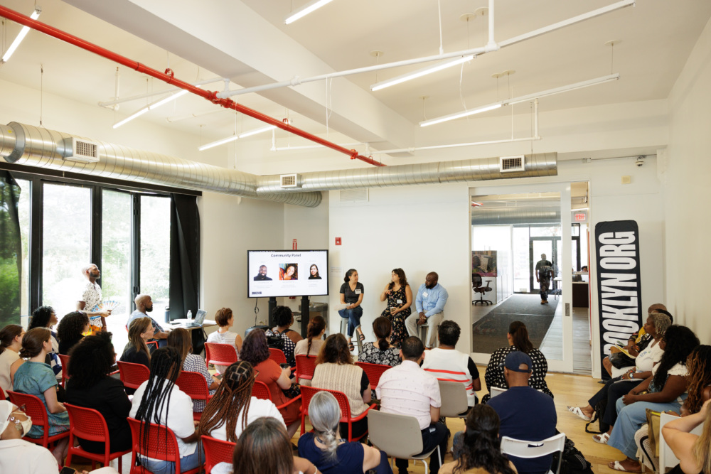 A group of people seated in a room attend a presentation. Three individuals stand near a screen at the front displaying portraits and names, while a large banner reads "BROOKLYN.ORG" to the right.