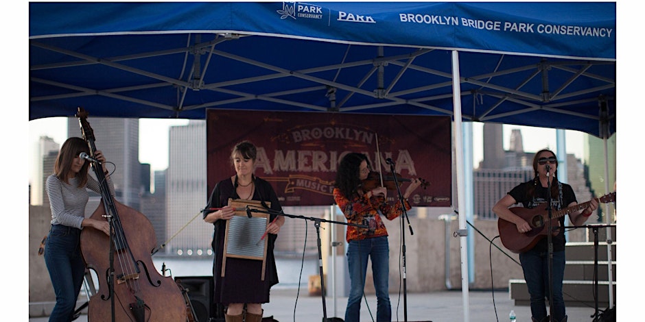 A four-member band performs on an outdoor stage under a blue tent with "Brooklyn Americana Music" signage in the background. The musicians play double bass, washboard, violin, and guitar.