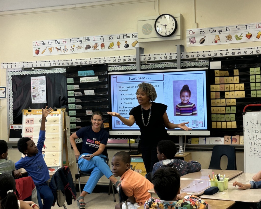 A teacher stands in front of a classroom full of students, speaking and gesturing with her hands. Another adult sits on a chair to her left. A large monitor displays information behind them.