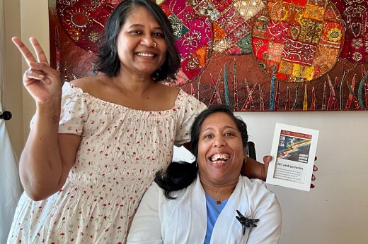 Two women smiling indoors; one standing, flashing a peace sign, and the other seated, holding a book titled "Art and Activism." The background features a colorful, textured wall art piece.