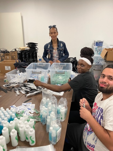 Three people are assembling hygiene kits at a table, organizing items like bottles and plastic bags in a room with stacked boxes and electronic equipment.