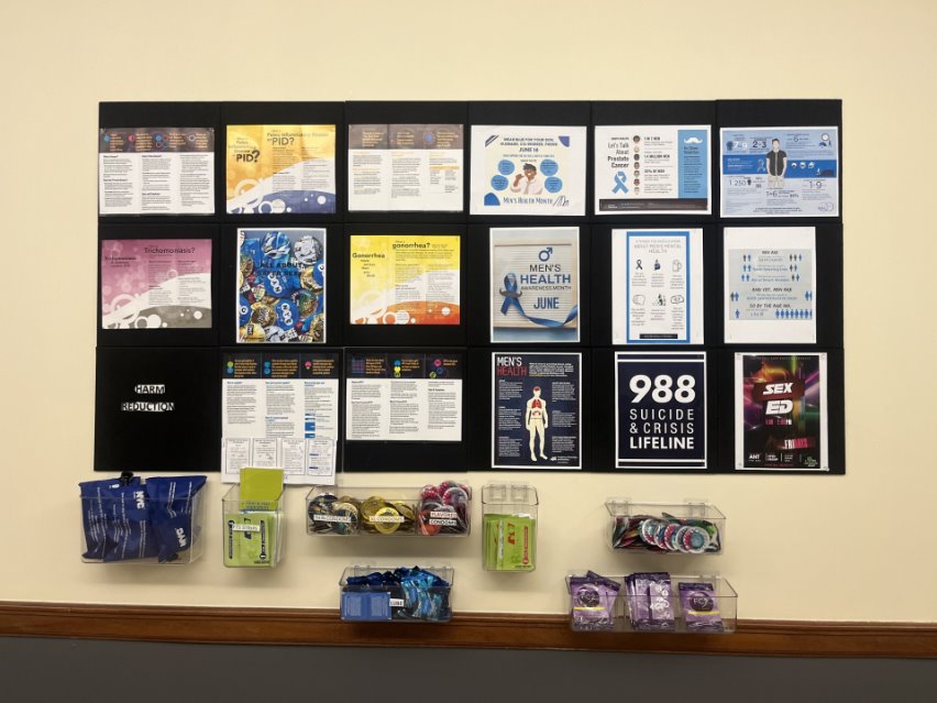 A display board with various health brochures, informational posters, and pamphlets about mental health, men's health, and safe sex, along with a selection of free condoms and lube packets.