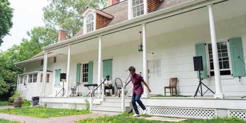 A person walks down the steps of a large white house with light blue shutters. A piano and speakers are set up on the porch, and another individual is seated playing the piano.