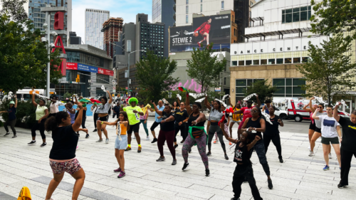 A group of people participate in an outdoor dance exercise class on a city street, led by an instructor facing them. Buildings and trees surround the area.