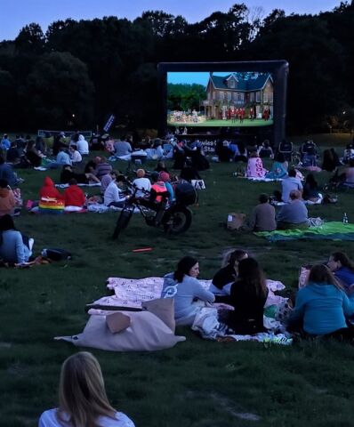 A crowd of people seated on blankets are watching an outdoor movie on a large inflatable screen in a park during the evening.