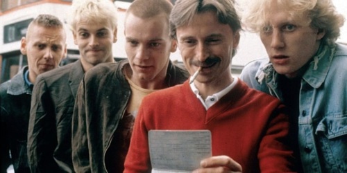 Five men standing closely together. One man, in a red sweater, is holding a document and smiling with a cigarette in his mouth. The others are looking at the document curiously.