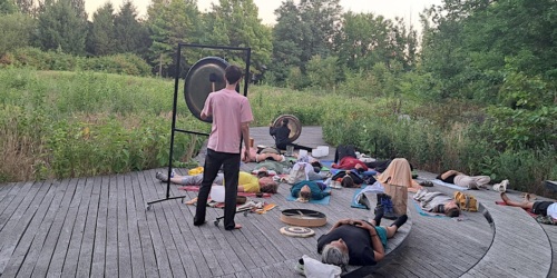 A group of people lying on mats outdoors in a wooded area, with one person standing and playing a gong.