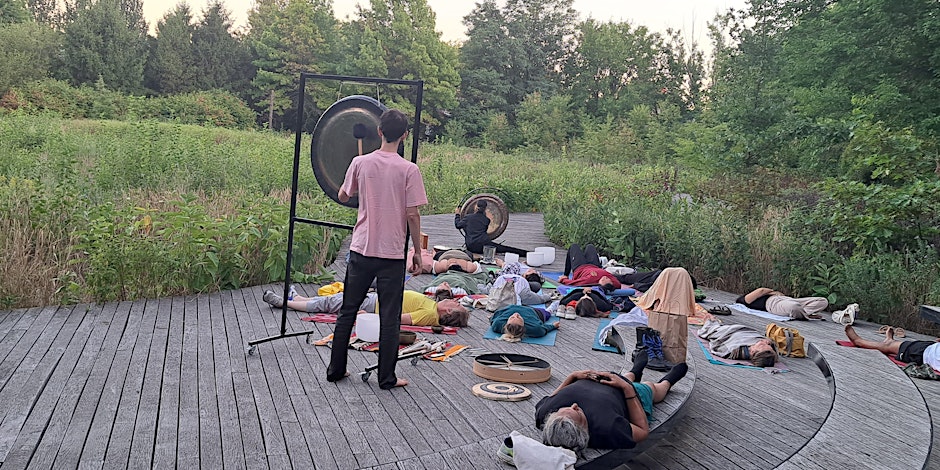 A group of people lying on mats outdoors in a wooded area, with one person standing and playing a gong.