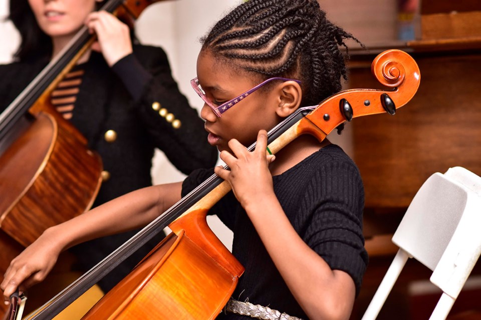 A child wearing glasses and a black shirt plays the cello. Another person playing a cello is partially visible in the background.