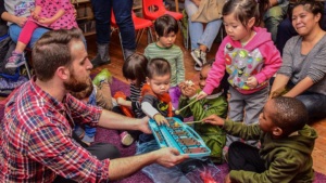 A group of young children and an adult engage with a blue toy boat during a group activity in a room with wooden floors. The children appear curious and interactive.