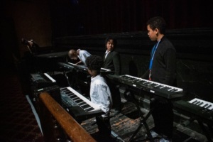 Four children stand near electronic keyboards on a dimly lit stage, engaged in conversation and activity.