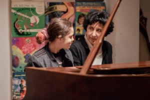 Two people sitting at a piano look at each other, with colorful posters on the wall behind them.