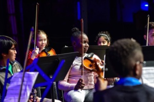 A group of children holding violins are seated in a dimly lit room, possibly preparing for a musical performance. Some have sheet music on stands in front of them.