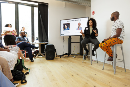 Two people are sitting on stools in front of a small audience, discussing a presentation titled "Partnership Modeling" displayed on a screen.