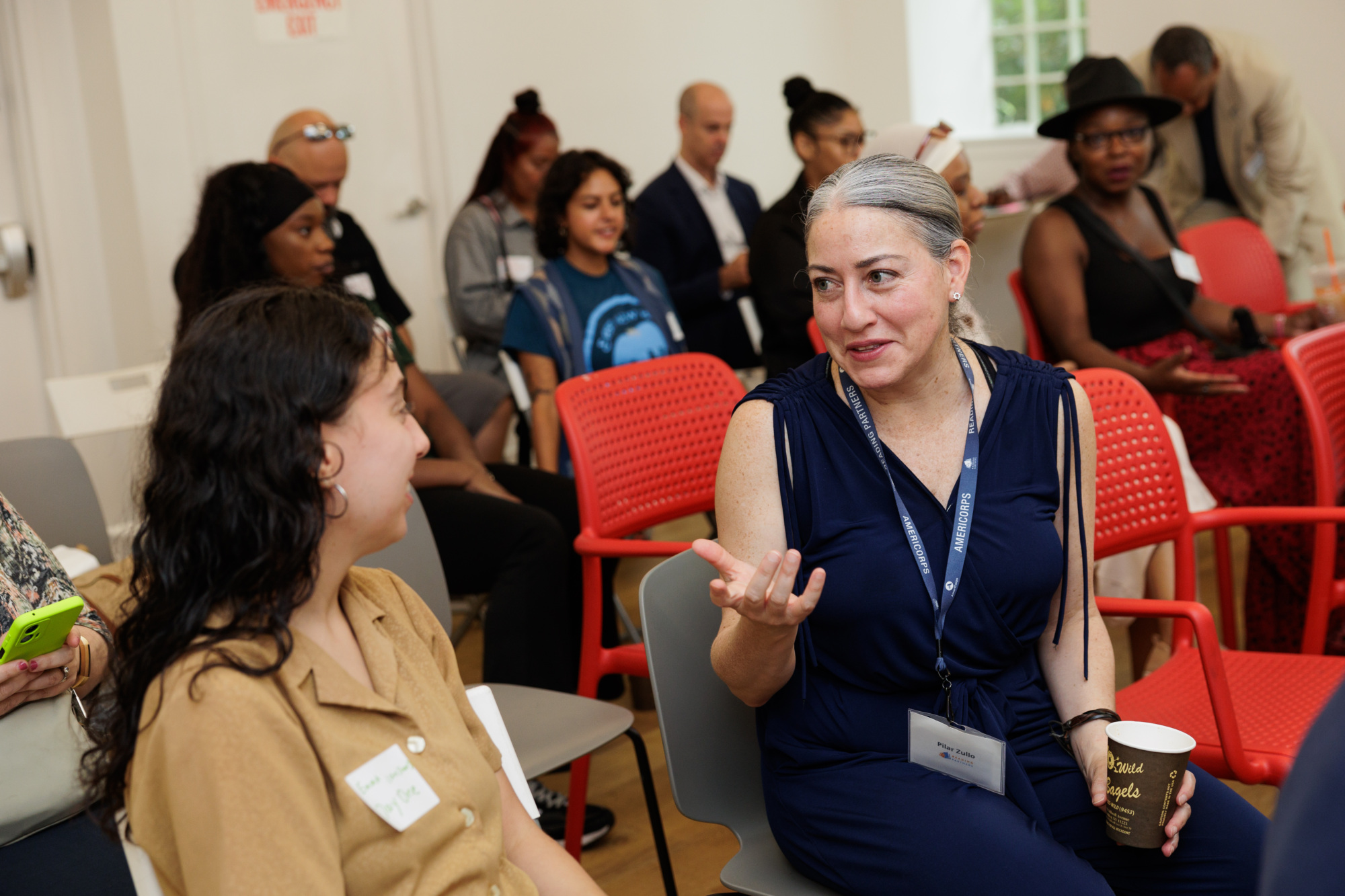 A group of people are seated in a room. In the foreground, two women are engaged in conversation; the woman on the right holds a cup, the woman on the left wears a brown shirt and has a name tag.