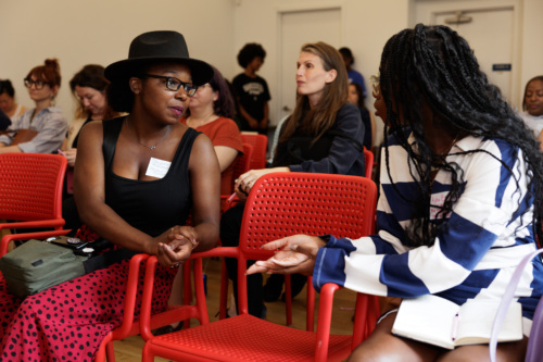 Two women seated in red chairs engage in conversation during a gathering in a room with several other attendees in the background.