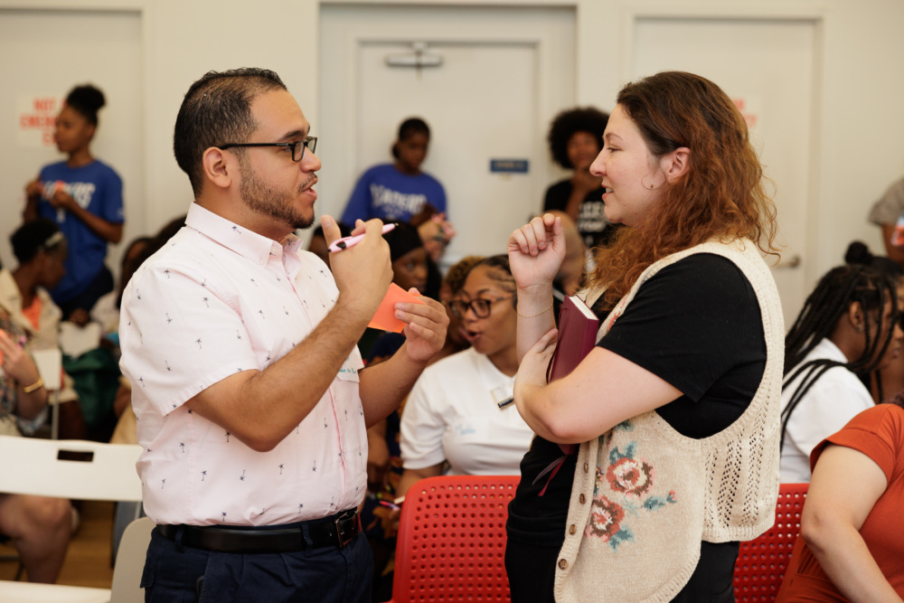 Two adults engage in conversation in a room full of people. One is holding a pen and the other a notebook. Other attendees can be seen talking and sitting in the background.