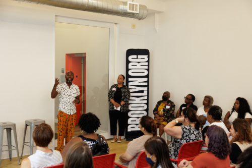 A person is speaking to a seated audience in a room. Another person stands nearby. A vertical sign reads "BROOKLYN.ORG".