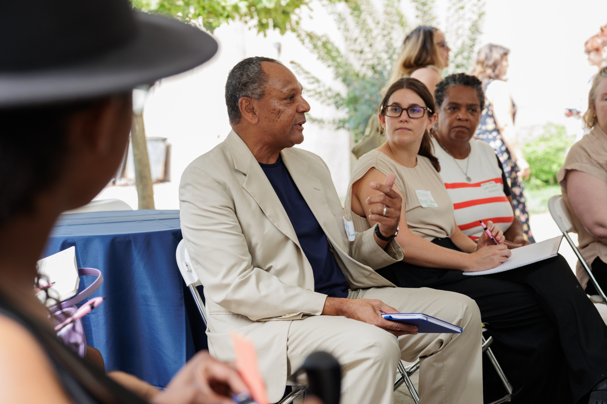 A group of people attending an outdoor meeting seated in a circle. A man in a cream-colored suit speaks while others listen and take notes.