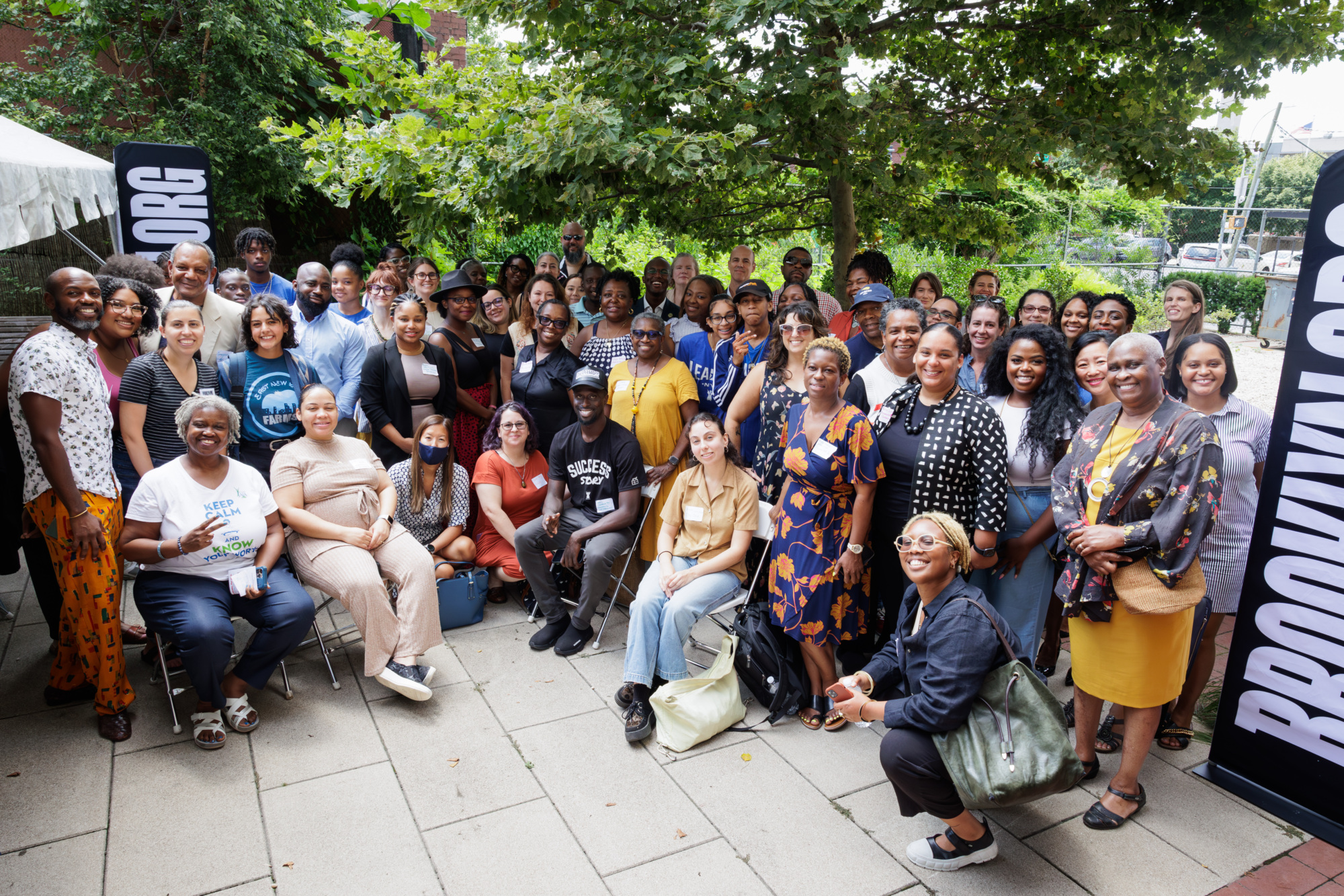 A large, diverse group of people posed together outdoors, standing and sitting on a paved area with greenery and "Brooklyn.org" banners in the background.