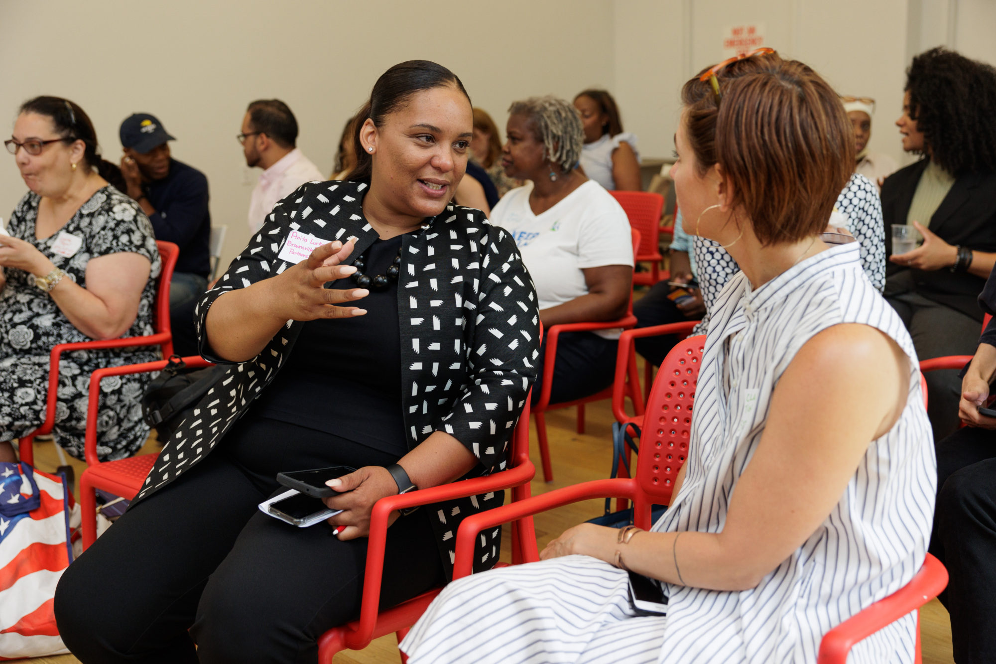 Two women engaged in conversation while sitting on red chairs in a room with other people. One woman is gesturing with her hand, and both have mobile phones in their laps.