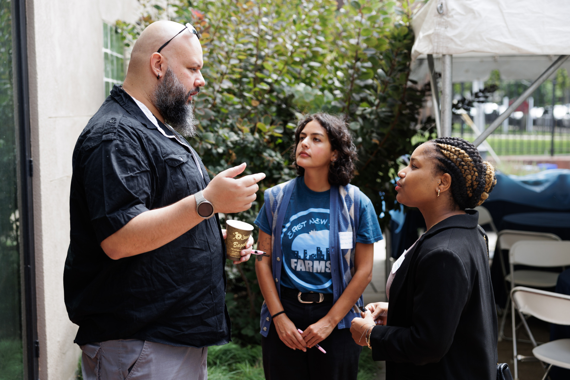 Three people having a discussion outdoors, with one man holding a coffee cup while two women listen attentively. They are under a white tent and greenery is in the background.