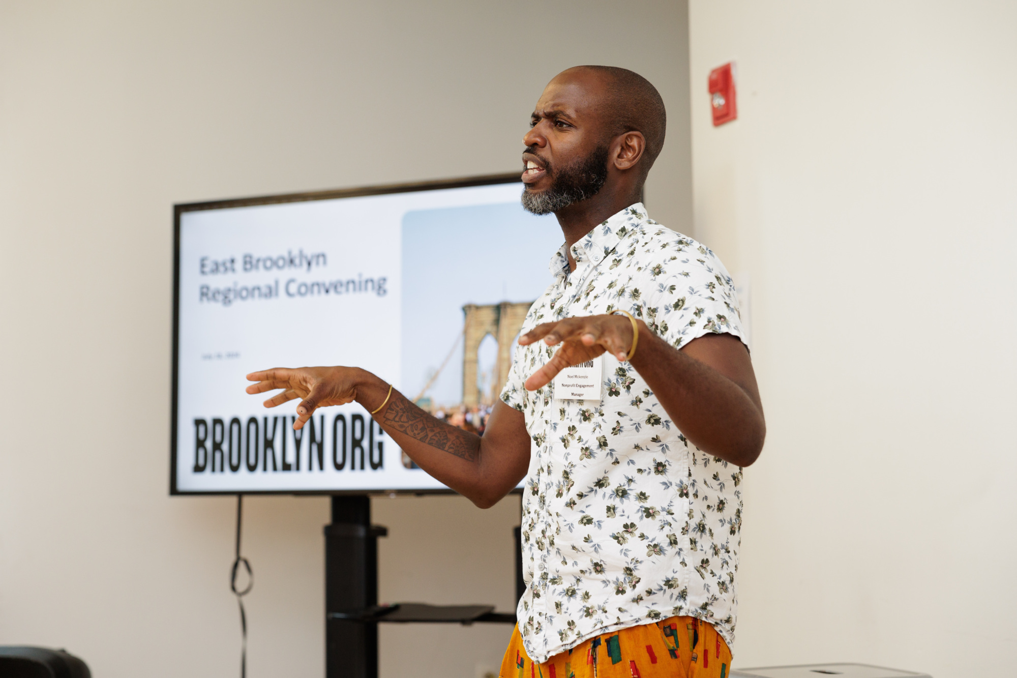 A person stands speaking in front of a screen displaying "East Brooklyn Regional Convening" and an image of the Brooklyn Bridge.