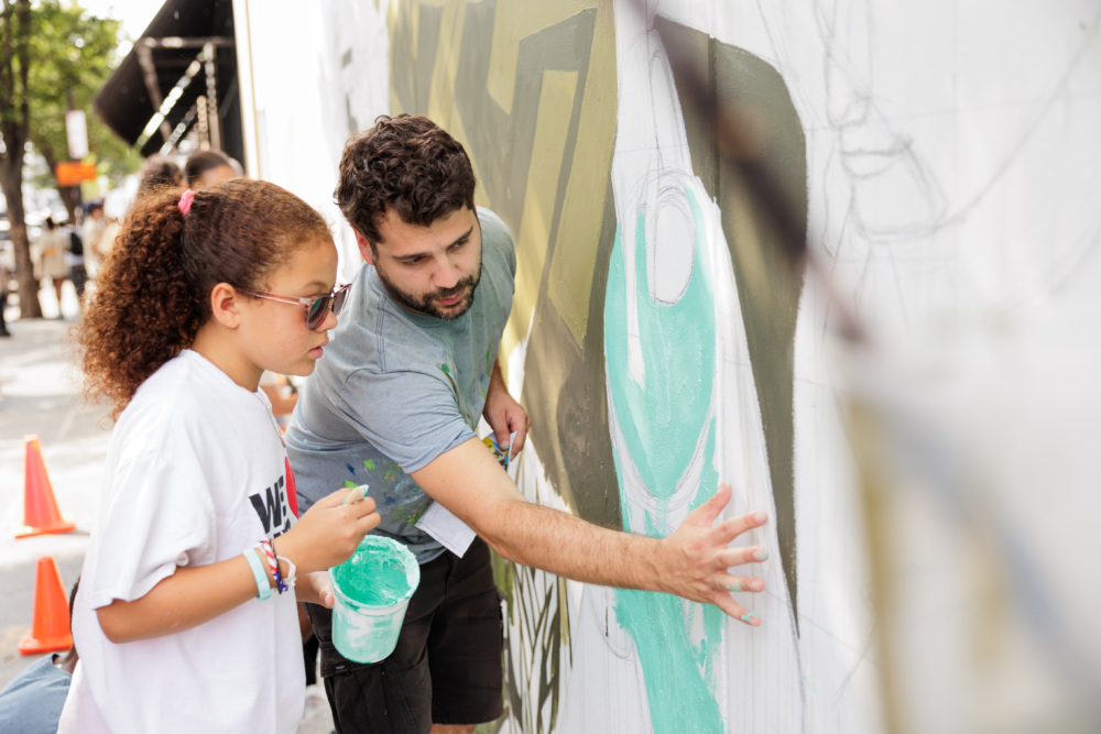 Two people, one holding a paint bucket, collaborate on painting a mural on an outdoor wall. Orange cones are visible in the background.