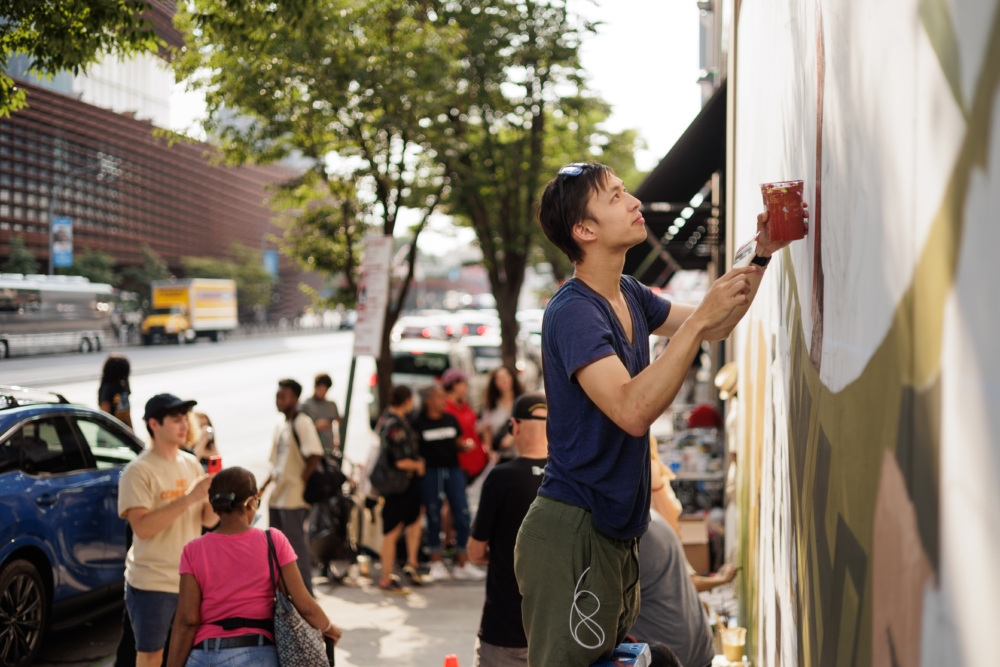 A person paints a mural on a wall along a busy street with a group of people nearby.