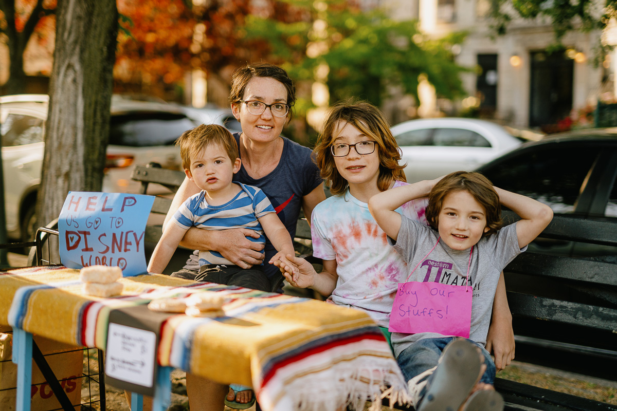 An adult and three children are seated at a small outdoor stand with a sign reading "Help us go to Disney World." The stand displays items for sale on a striped tablecloth.