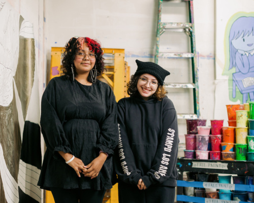 Two individuals in black clothing stand in an art studio, with paint cans, a ladder, and artwork in the background.