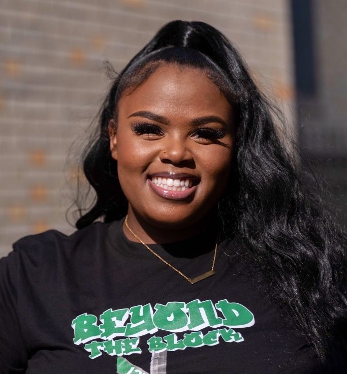 Woman smiling outside, wearing a black T-shirt with "BEYOND THE BLOCK" printed in green and white letters. She has long black hair styled in a high ponytail.